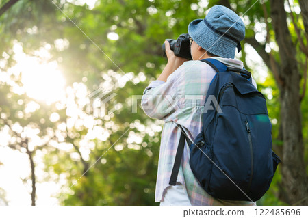 Portrait of Asian mature woman holding a camera and backpack behind her back, an Asia active senior woman enjoying nature in park. Standing on a trail in a forest outdoors. Enjoying active travel trip 122485696