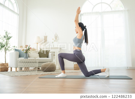 Young Asian woman in sportswear doing yoga in her home or house for health, sports and wellness, Enjoying Domestic Fitness Training And Healthy Lifestyle, Dressed In Stylish Sportswear And Smiling 122486233