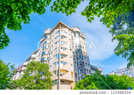 A residential block of flats in Belgrade, Serbia is framed by green trees and a bright blue sky. The architectural design features rounded edges, providing a unique look to the area. A residential block of flats in Belgrade, Serbia is framed by green trees and a bright blue sky. The architectural design features rounded edges, providing a unique look to the area. 122486354