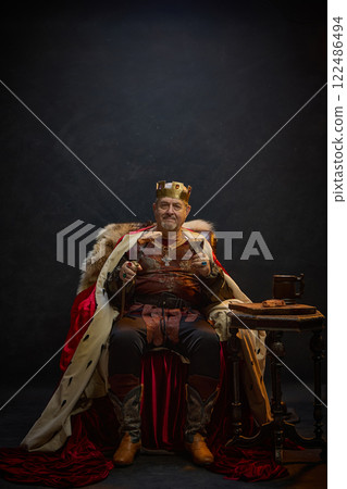 Senior man, medieval king in period attire sitting on throne with meat on sword and glass of wine against black studio background Senior man, medieval king in period attire sitting on throne with meat on sword and glass of wine against black studio background 122486494