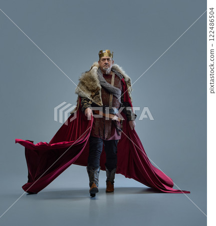 Elderly focused man, medieval king walks forward with determination, cloak flowing behind against grey studio background. Progress, leadership. 122486504