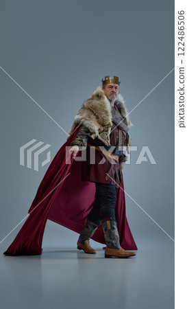 Elderly focused man, medieval king walks forward with determination, cloak flowing behind against grey studio background. 122486506