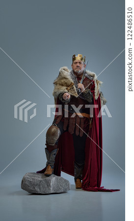 Choice. Full-length portrait of medieval king with crown and sword, standing with determination and pointing forwards against grey studio background Choice. Full-length portrait of medieval king with crown and sword, standing with determination and pointing forwards against grey studio background 122486510