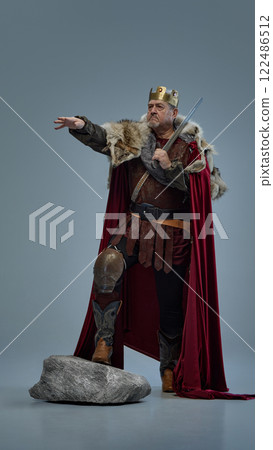 Full-length portrait of senior man as medieval king, in crown with sword, standing on stone and pointing forward against grey studio background 122486512