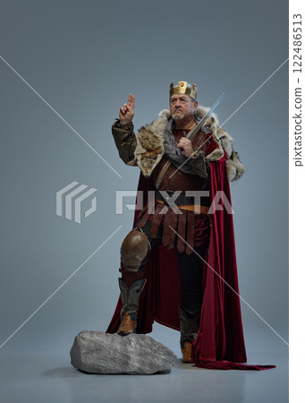 Senior serious man as medieval king standing on stone with sword and gesturing with one hand raised against grey studio background. Hope 122486513