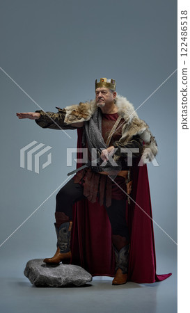 Commanding medieval king on rock, pointing forward with authority against grey studio background. 122486518