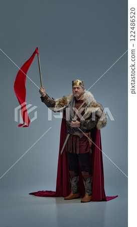 Powerful medieval king holding red flag, standing with determination against grey studio background. Powerful medieval king holding red flag, standing with determination against grey studio background. 122486520