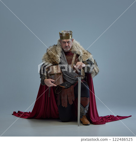 Elderly man, medieval king with crown and fur cloak holding metal helmet and sitting on one knee against grey studio background. Elderly man, medieval king with crown and fur cloak holding metal helmet and sitting on one knee against grey studio background. 122486525