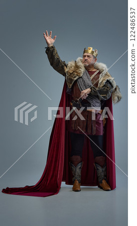 Serious senior man as medieval king in red cape and fir-lined armor, raising hand in commanding gesture against grey studio background Serious senior man as medieval king in red cape and fir-lined armor, raising hand in commanding gesture against grey studio background 122486537