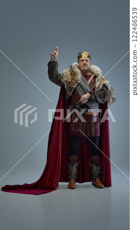 Portrait of elderly man as medieval king in red cape and fur-lined armor, raising hand in gesture against grey studio background 122486539