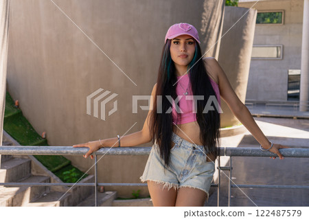 Young millennial smiling latina woman with a pink cap and long hair leaning on a railing 122487579