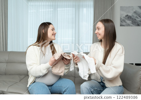 Young pregnant woman and her female friend looking at baby clothes while sitting on a couch. Concept of motherhood, friendship, preparation, excitement, and bonding. Young pregnant woman and her female friend looking at baby clothes while sitting on a couch. Concept of motherhood, friendship, preparation, excitement, and bonding. 122489386