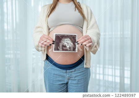 Pregnant woman holding ultrasound scan photo of a fetus in front of her belly in third trimester at home. Concept of pregnancy, sonography. Maternity prenatal care Pregnant woman holding ultrasound scan photo of a fetus in front of her belly in third trimester at home. Concept of pregnancy, sonography. Maternity prenatal care 122489397