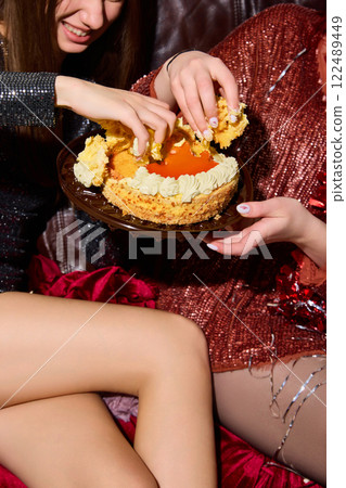 Two women eating cake with hands, laughing, sitting on brown sofa, dressed in black and red dresses, surrounded by messy party decorations. Two women eating cake with hands, laughing, sitting on brown sofa, dressed in black and red dresses, surrounded by messy party decorations. 122489449
