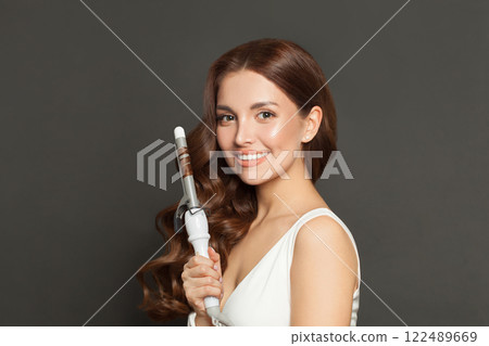Glamurous brown haired woman with shiny long curly hairstyle showing curling iron.  122489669
