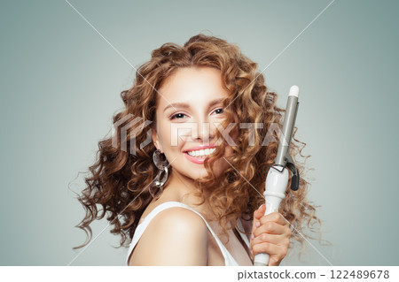Laughing brown haired woman with shiny long curly hairstyle showing curling iron.  122489678