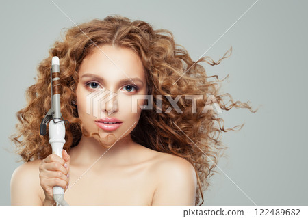 Perfect woman with curly hairstyle using hot curling tongs iron, indoor fashion portrait.  122489682