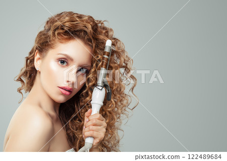 Pretty woman with curly hairstyle using hot curling tongs iron, indoor fashion portrait.  122489684