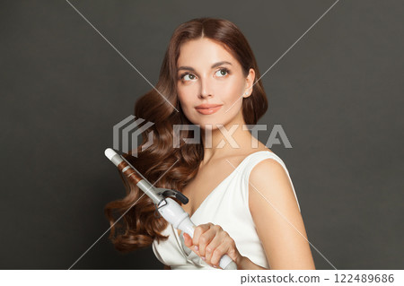 Attractive woman with curly hairstyle using hot curling tongs iron, indoor fashion portrait. 122489686