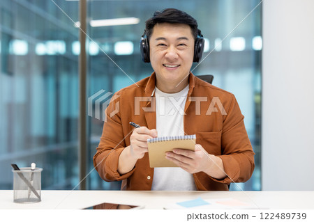 Webcam view, Asian smiling man in headphones looking at camera, listening and watching training webinar, writing in notebook. Man inside office at workplace at online business meeting conference 122489739