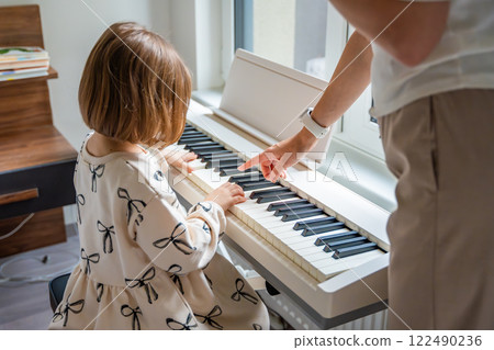 Home lesson with teacher on music for girl on white piano synthesizer. Concept of early development: learning music enhances motor skills, hearing, intelligence, fostering creativity from childhood Home lesson with teacher on music for girl on white piano synthesizer. Concept of early development: learning music enhances motor skills, hearing, intelligence, fostering creativity from childhood 122490236