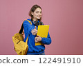 Front view of schoolgirl standing, looking at camera, smiling. Beautiful young teenage female with earphones and rucksack, showing super. Concept of education and youth. 122491910