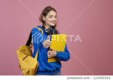 Front view of schoolgirl standing, looking at camera, smiling. Beautiful young teenage female with earphones and rucksack, showing super. Concept of education and youth. 122491910