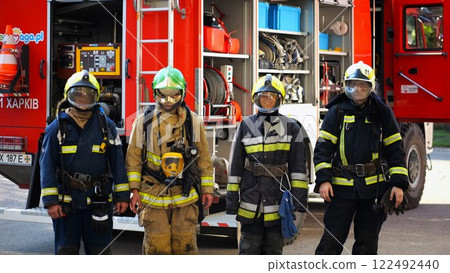 Portrait of male and female firefighters in helmets and protective uniforms standing near big red car. Young confident fireman and firewoman in full equipment posing against background of fire engine Portrait of male and female firefighters in helmets and protective uniforms standing near big red car. Young confident fireman and firewoman in full equipment posing against background of fire engine 122492440