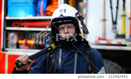 Portrait of confident male fireguard with axe standing near a fire engine. Young fireman with dirty face looking at camera in full equipment after fire against the background of big red truck 122492441