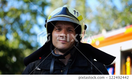 Portrait of young confident fireguard in protective uniform against background of a fire truck. Male firefighter in full equipment looking into camera with positive emotions near big red car. Close up 122492442