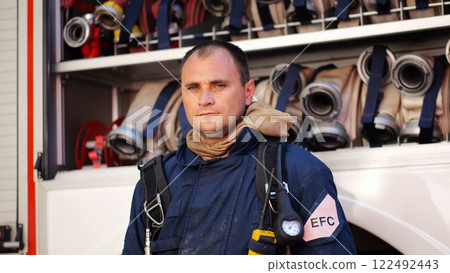 Portrait of confident male firefighter standing near a fire engine. Young fireman in protective uniform posing against the background of big red truck. Concept of saving lives and heroic profession Portrait of confident male firefighter standing near a fire engine. Young fireman in protective uniform posing against the background of big red truck. Concept of saving lives and heroic profession 122492443