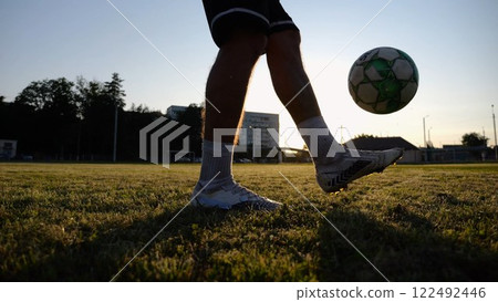 Legs of young man kicking ball at green field. Male feet of professional footballer juggling soccer ball on stadium at sunset. Sportsman practicing tricks outdoor. Concept of a freestyle football 122492446
