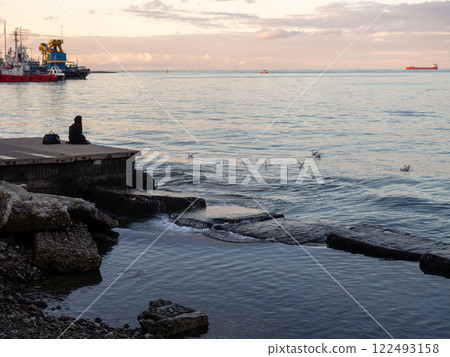 A lonely girl sits on a pier in the port. Sitting on the edge of the water. 122493158