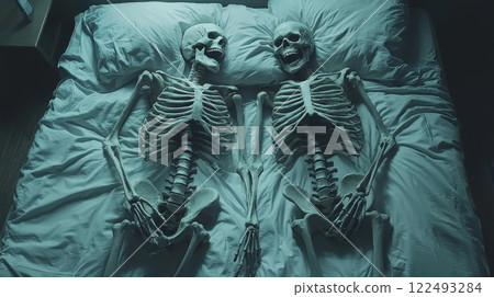 Two human skeletons lying side by side on a bed in a dimly lit room reflecting eerie and mysterious atmosphere of halloween night 122493284