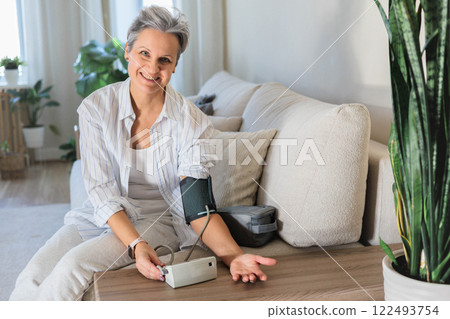Mature beautiful middle-aged woman with gray hair sits on the sofa and measures blood pressure  122493754