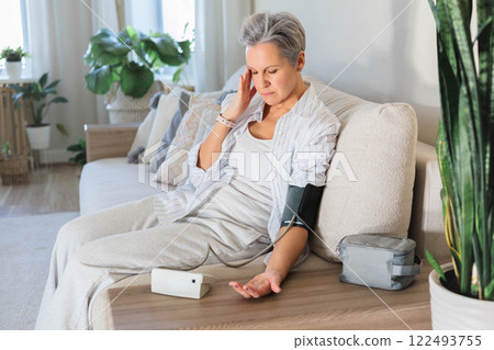 An elderly woman with gray hair sits on the couch and measures blood pressure with a tonometer. An elderly woman with gray hair sits on the couch and measures blood pressure with a tonometer. 122493755