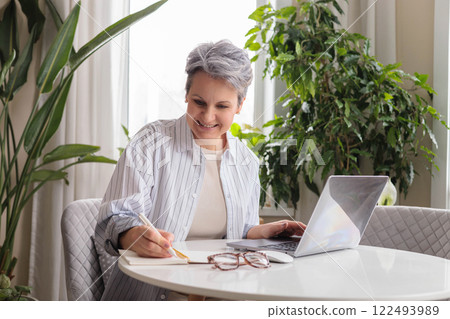 Smiling stylish mature middle aged woman sitting at table with laptop, portrait. Smiling stylish mature middle aged woman sitting at table with laptop, portrait. 122493989
