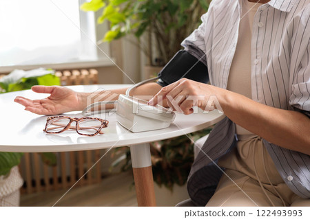 A woman in a blue shirt measures her blood pressure with a tonometer on a white table. A woman in a blue shirt measures her blood pressure with a tonometer on a white table. 122493993