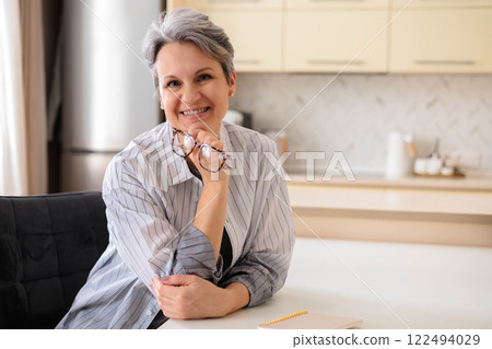 A beautiful elderly woman pensioner with gray hair sits at the table at home in her kitchen. A beautiful elderly woman pensioner with gray hair sits at the table at home in her kitchen. 122494029