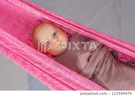 A 3-month-old baby peacefully falls asleep in a soft muslin swaddle while cradled in a cozy hammock. A serene and tender moment of comfort and relaxation. Baby care, rest, and tranquility concept 122494474