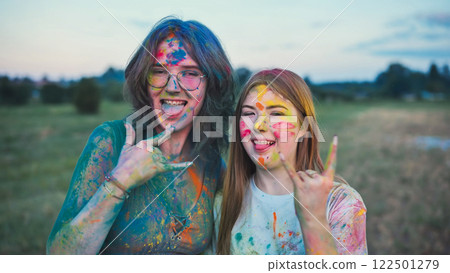 Two young women, covered in colorful powder paint, are enjoying the holi festival, sticking their tongues out and making the rock and roll hand sign Two young women, covered in colorful powder paint, are enjoying the holi festival, sticking their tongues out and making the rock and roll hand sign 122501279