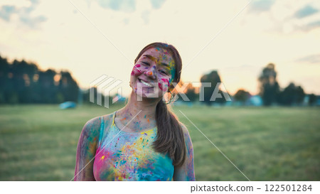 Happy young woman covered in vivid colors, smiling during the holi festival in a field at sunset Happy young woman covered in vivid colors, smiling during the holi festival in a field at sunset 122501284