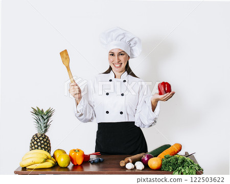 Woman chef holding a ladle on a table with vegetables, A smiling woman chef holding a ladle and vegetables, portrait of a woman chef holding a ladle on isolated background Woman chef holding a ladle on a table with vegetables, A smiling woman chef holding a ladle and vegetables, portrait of a woman chef holding a ladle on isolated background 122503622
