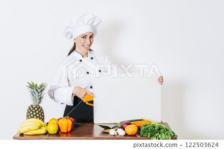 Chef woman with vegetables at table holding blank menu. Girl chef in the kitchen showing a blank board, Beautiful woman chef with table of vegetables holding a blank board Chef woman with vegetables at table holding blank menu. Girl chef in the kitchen showing a blank board, Beautiful woman chef with table of vegetables holding a blank board 122503624