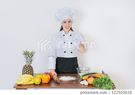 Chef woman holding bread rolling pin with thumb up, Smiling chef woman holding rolling pin on vegetable table, Concept of chef woman holding bread rolling pin 122503628