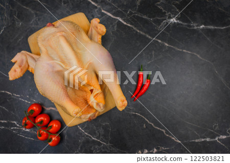 Close-up of a chicken, butchered and plucked, lying on a wooden plank against a background of black marble, surrounded by cherry tomatoes and hot red peppers, with space for text on the right. The 122503821