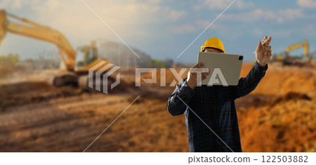 engineer working at site of a large building man wearing a yellow hard hat is looking at a tablet while standing in front engineer working at site of a large building man wearing a yellow hard hat is looking at a tablet while standing in front 122503882