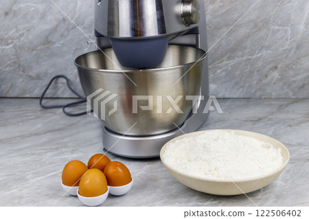 Modern kitchen machine and ingredients for preparing dough (eggs and flour) on a kitchen table Modern kitchen machine and ingredients for preparing dough (eggs and flour) on a kitchen table 122506402
