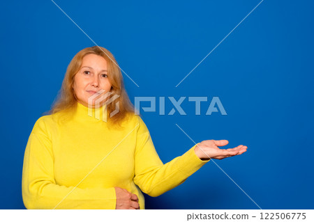 Caucasian woman in her 40s wearing a yellow sweater pointing at something on open palm, demonstrating invisible object, free space for mockup, blue background, panorama. Caucasian woman in her 40s wearing a yellow sweater pointing at something on open palm, demonstrating invisible object, free space for mockup, blue background, panorama. 122506775
