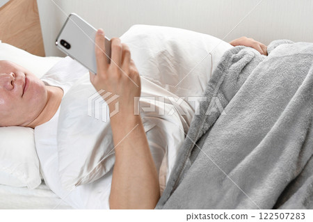 A man smiling while lying in bed and using his smartphone A man smiling while lying in bed and using his smartphone 122507283